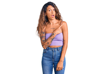 Young hispanic woman with tattoo wearing casual clothes surprised pointing with finger to the side, open mouth amazed expression.