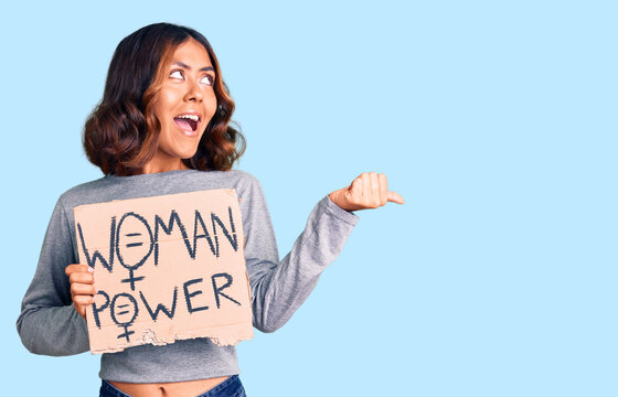 Young beautiful mixed race woman holding woman power banner pointing thumb up to the side smiling happy with open mouth