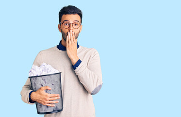 Young handsome hispanic man holding paper bin full of crumpled papers covering mouth with hand, shocked and afraid for mistake. surprised expression