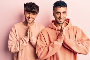 Young gay couple wearing casual clothes praying with hands together asking for forgiveness smiling confident.