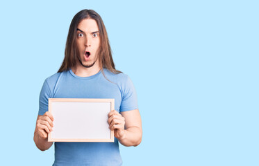Young adult man with long hair holding empty white chalkboard scared and amazed with open mouth for surprise, disbelief face