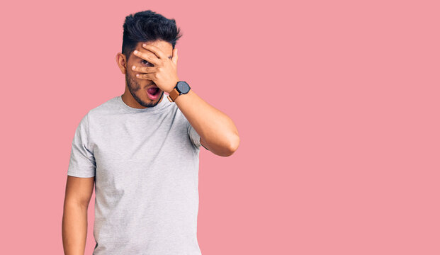 Handsome latin american young man wearing casual tshirt peeking in shock covering face and eyes with hand, looking through fingers with embarrassed expression.