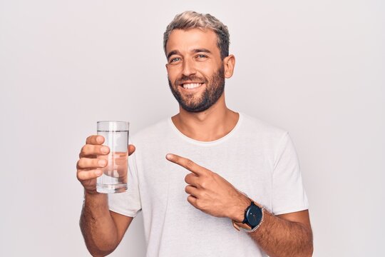 Handsome blond man with beard drinking glass of water to refreshment over white background smiling happy pointing with hand and finger
