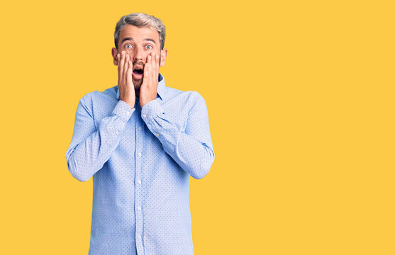 Young handsome blond man wearing elegant shirt afraid and shocked, surprise and amazed expression with hands on face