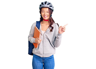 Young beautiful chinese girl wearing student backpack and bike helmet holding book smiling happy pointing with hand and finger to the side