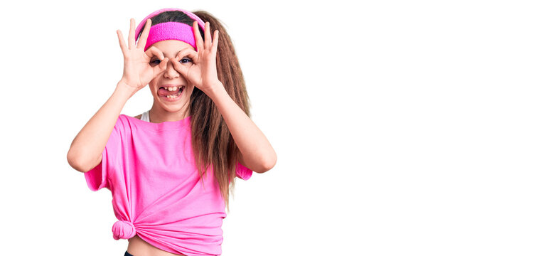 Cute hispanic child girl wearing gym clothes and using headphones doing ok gesture like binoculars sticking tongue out, eyes looking through fingers. crazy expression.