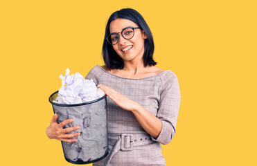 Young beautiful latin woman holding paper bin full of crumpled papers looking positive and happy standing and smiling with a confident smile showing teeth