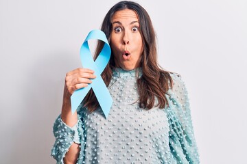 Young beautiful brunette woman holding blue ribbon symbol over isolated white background scared and amazed with open mouth for surprise, disbelief face