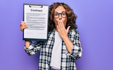 Middle age brunette business woman wearing glasses holding clipboard with contract paper sheet covering mouth with hand, shocked and afraid for mistake. Surprised expression