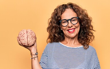 Middle age brunette woman holding brain as mental care and memory health over pink background...