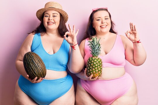 Young plus size twins wearing bikini holding melon and pineapple doing ok sign with fingers, smiling friendly gesturing excellent symbol