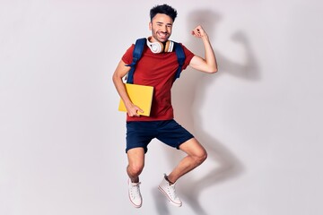 Young handsome hispanic student man wearing backpack smiling happy. Jumping with smile on face holding book using headphones. Celebrating with fist up over isolated white background.