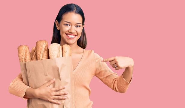 Young beautiful latin girl holding paper bag with bread pointing finger to one self smiling happy and proud