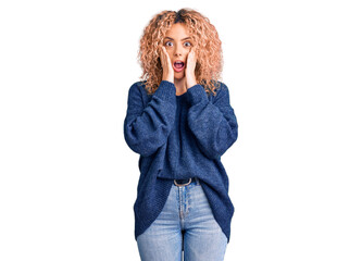 Young blonde woman with curly hair wearing casual winter sweater afraid and shocked, surprise and amazed expression with hands on face