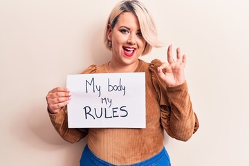 Fototapeta na wymiar Blonde plus size woman asking for women rights holding paper with my body my rules message doing ok sign with fingers, smiling friendly gesturing excellent symbol