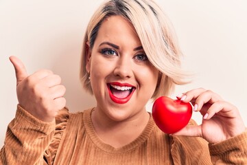 Young beautiful blonde plus size woman holding plastic heart over isolated white background pointing thumb up to the side smiling happy with open mouth