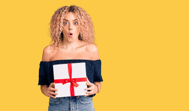 Young blonde woman with curly hair holding gift scared and amazed with open mouth for surprise, disbelief face