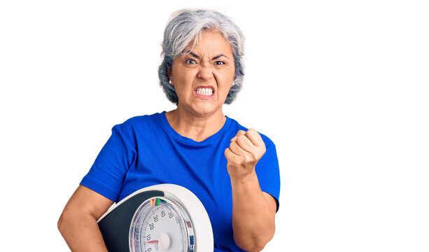 Senior woman with gray hair holding weight machine to balance weight loss annoyed and frustrated shouting with anger, yelling crazy with anger and hand raised