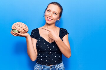 Young beautiful blonde woman holding brain smiling happy pointing with hand and finger
