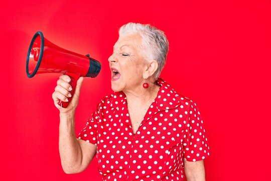 Senior beautiful grey-haired woman screaming using megaphone over isolated red background
