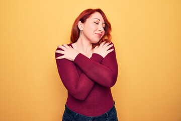 Young beautiful redhead woman wearing casual sweater standing over yellow background hugging...