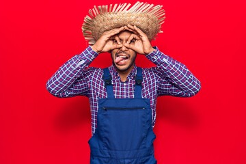Young latin man wearing farmer hat and apron doing ok gesture like binoculars sticking tongue out, eyes looking through fingers. crazy expression.
