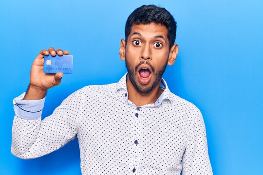 Young latin man holding credit card scared and amazed with open mouth for surprise, disbelief face