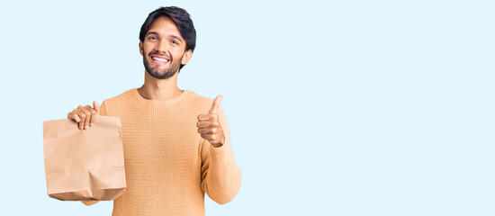 Handsome hispanic man holding take away paper bag smiling happy and positive, thumb up doing excellent and approval sign
