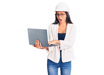 Young beautiful latin girl wearing architect hardhat holding laptop scared and amazed with open mouth for surprise, disbelief face