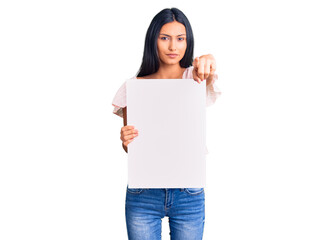 Young beautiful latin girl holding blank empty banner pointing with finger to the camera and to you, confident gesture looking serious