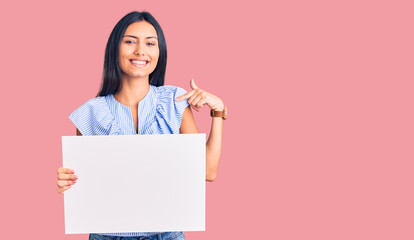 Fototapeta premium Young beautiful latin girl holding blank empty banner pointing finger to one self smiling happy and proud