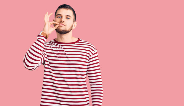 Young handsome man wearing striped sweater mouth and lips shut as zip with fingers. secret and silent, taboo talking