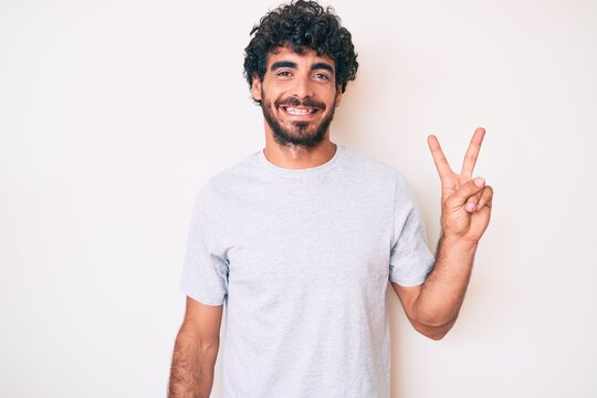 Handsome young man with curly hair and bear wearing casual tshirt smiling with happy face winking at the camera doing victory sign. number two.