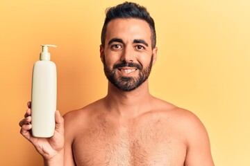 Young hispanic man standing shirtless holding sunscreen lotion looking positive and happy standing and smiling with a confident smile showing teeth