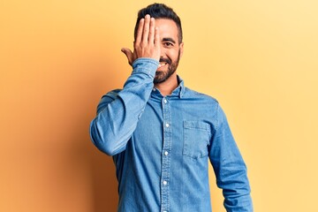 Young hispanic man wearing casual clothes covering one eye with hand, confident smile on face and surprise emotion.