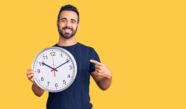 Young hispanic man holding big clock smiling happy pointing with hand and finger