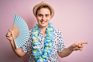 Young handsome redhead tourist man on vacation wearing hawaiian lei and hat using hand fan Smiling...