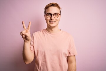 Naklejka premium Young handsome redhead man wearing casual t-shirt standing over isolated pink background smiling looking to the camera showing fingers doing victory sign. Number two.