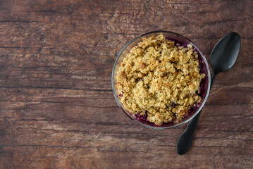 Top view of freshly baked blueberry crunch in a glass bowl on a rustic wood table, black spoon
