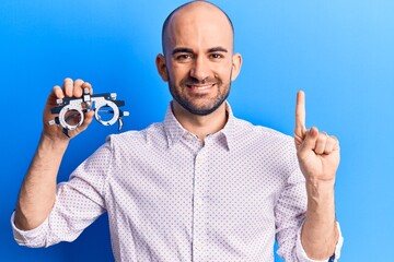 Young handsome bald man holding optometry glasses smiling with an idea or question pointing finger with happy face, number one