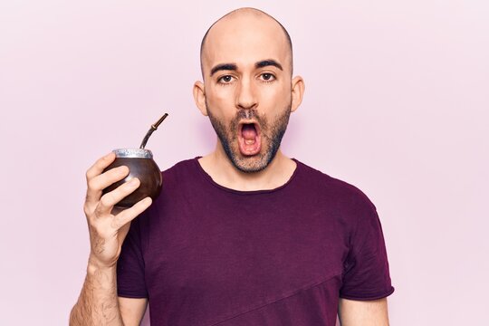Young handsome bald man drinking mate infusion beverage scared and amazed with open mouth for surprise, disbelief face
