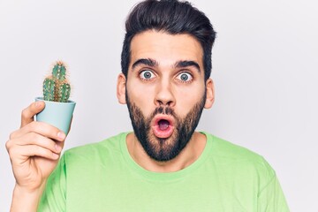 Young handsome man with beard holding small cactus pot scared and amazed with open mouth for...
