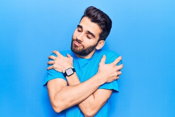 Young handsome man with beard wearing casual t-shirt hugging oneself happy and positive, smiling...