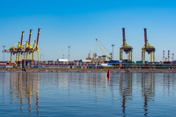 Naklejka premium Seaport. Water, port cranes and containers under a blue sky. Cargo transportation by water. Merchant fleet. Panorama of the port on a Sunny day.