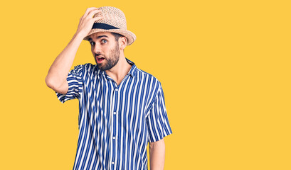 Young handsome man with beard wearing summer hat and striped shirt surprised with hand on head for mistake, remember error. forgot, bad memory concept.