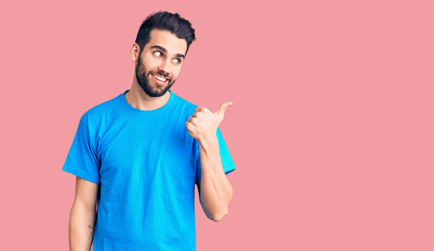 Young handsome man with beard wearing casual t-shirt smiling with happy face looking and pointing to the side with thumb up.
