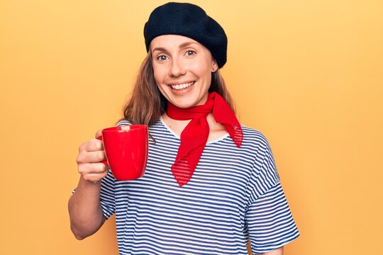 Young beautiful blonde woman wearing fashion beret drinking a cup of coffee looking positive and happy standing and smiling with a confident smile showing teeth