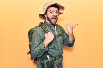 Young hispanic man wearing explorer hat and bakcpack pointing thumb up to the side smiling happy with open mouth