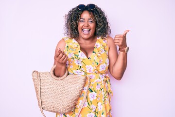 Young african american plus size woman wearing casual clothes holding summer wicker handbag pointing thumb up to the side smiling happy with open mouth