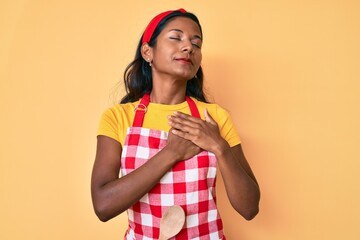 Young indian girl wearing baker apron smiling with hands on chest, eyes closed with grateful gesture on face. health concept.